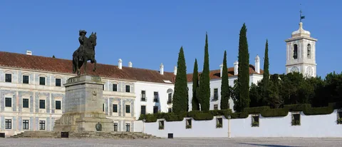 Hertugpalasset i Vila Viçosa med marmorfasade, rytterstatuen av kong João IV og sypresser under blå himmel