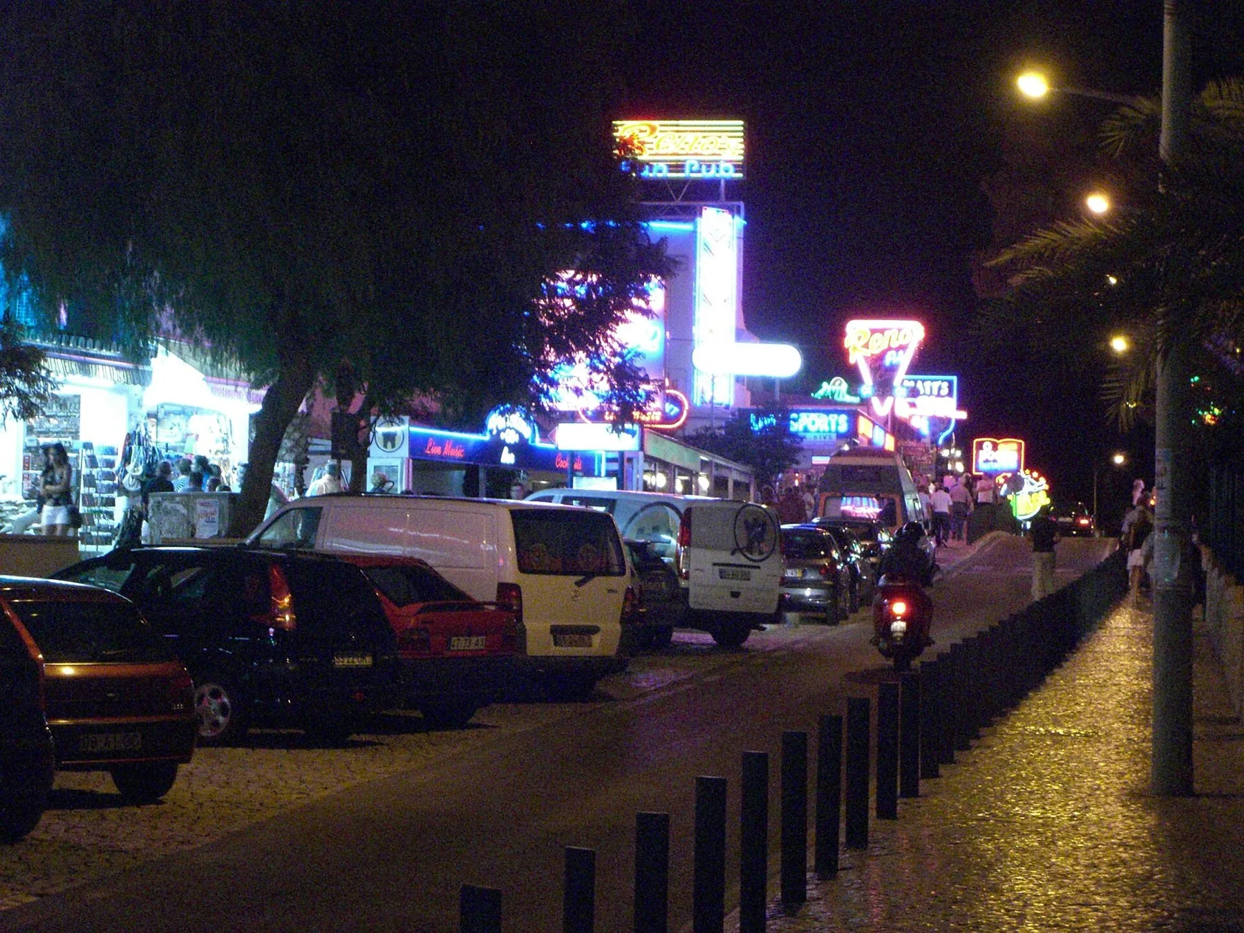 Neonskilt fra barer og klubber lyser opp The Strip i Albufeira om natten, med parkerte biler langs gata