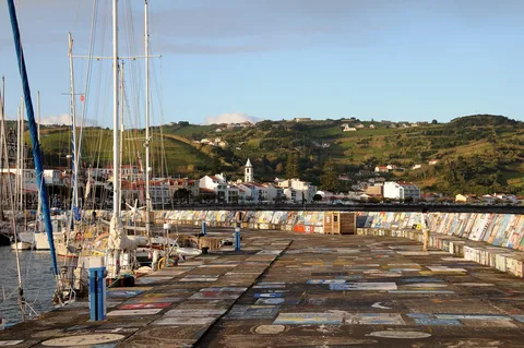 Seilbåter i Horta-marinaen og moloen dekket av seilernes malte minnebilder, med byen og grønne åser bak