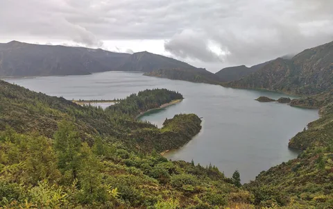 Kratersjøen Lagoa do Fogo med en skogkledd odde, omkranset av grønne kraterkanter under grå skyer