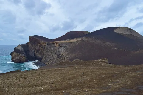 Brune askekjegler og lagdelte lavaklipper ved vulkanen Capelinhos der brenningene slår inn fra havet