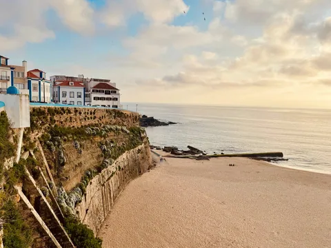 Sandstrand under klippen ved gamlebyen i Ericeira, med hvite hus på kanten og solnedgang over havet