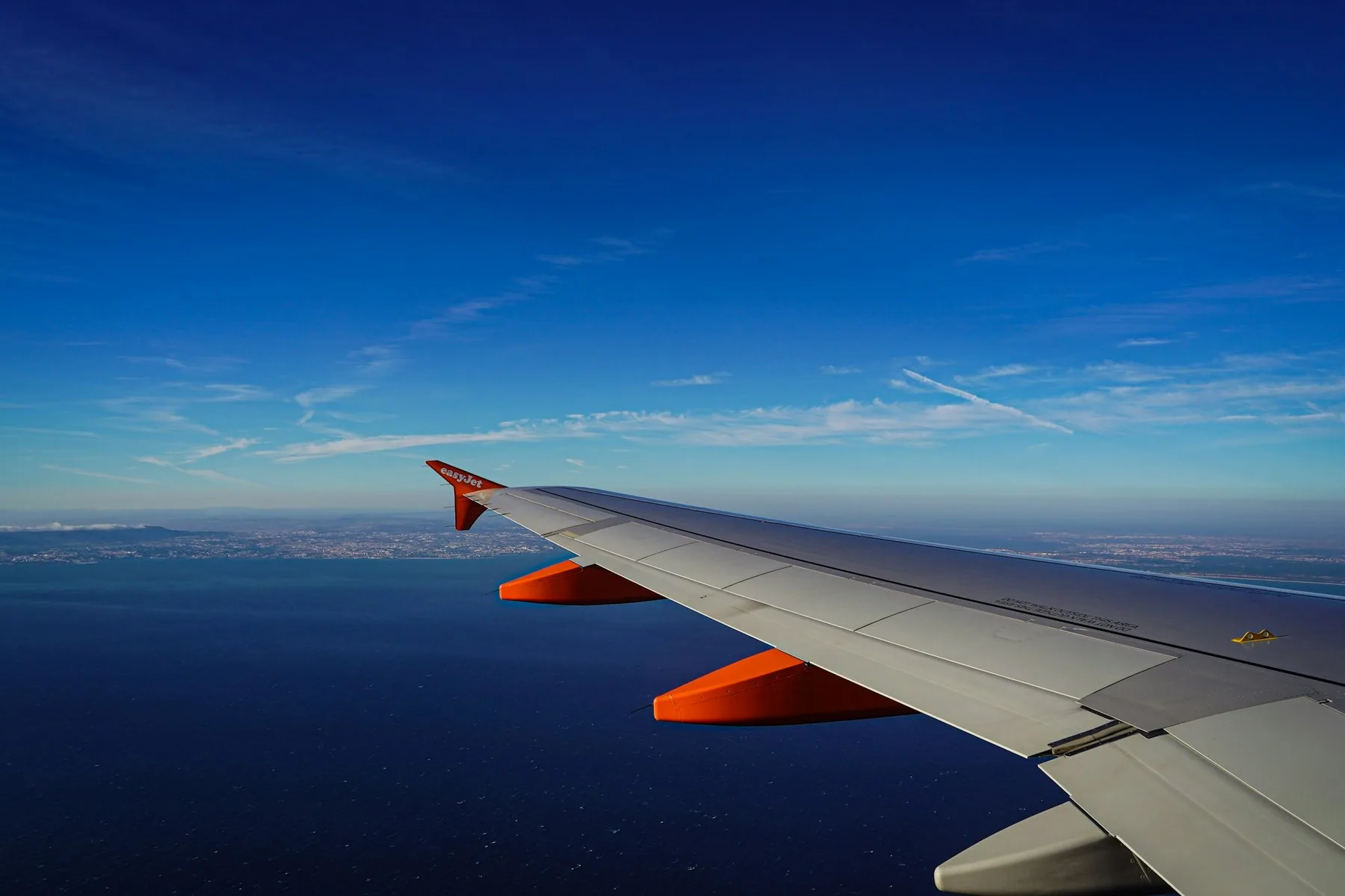 Flyvinge med oransje winglet over dypblått hav, med kysten ved Lisboa i det fjerne under klar himmel