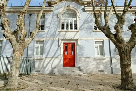 Lyseblå skolebygning med røde dører og buevindu bak nakne platantrær og steintrapp i Lisboa