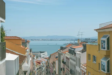 Bratt gate mellom bygårder med oransje tak i Estrela i Lisboa, med utsikt ned mot Tejo og havnekraner