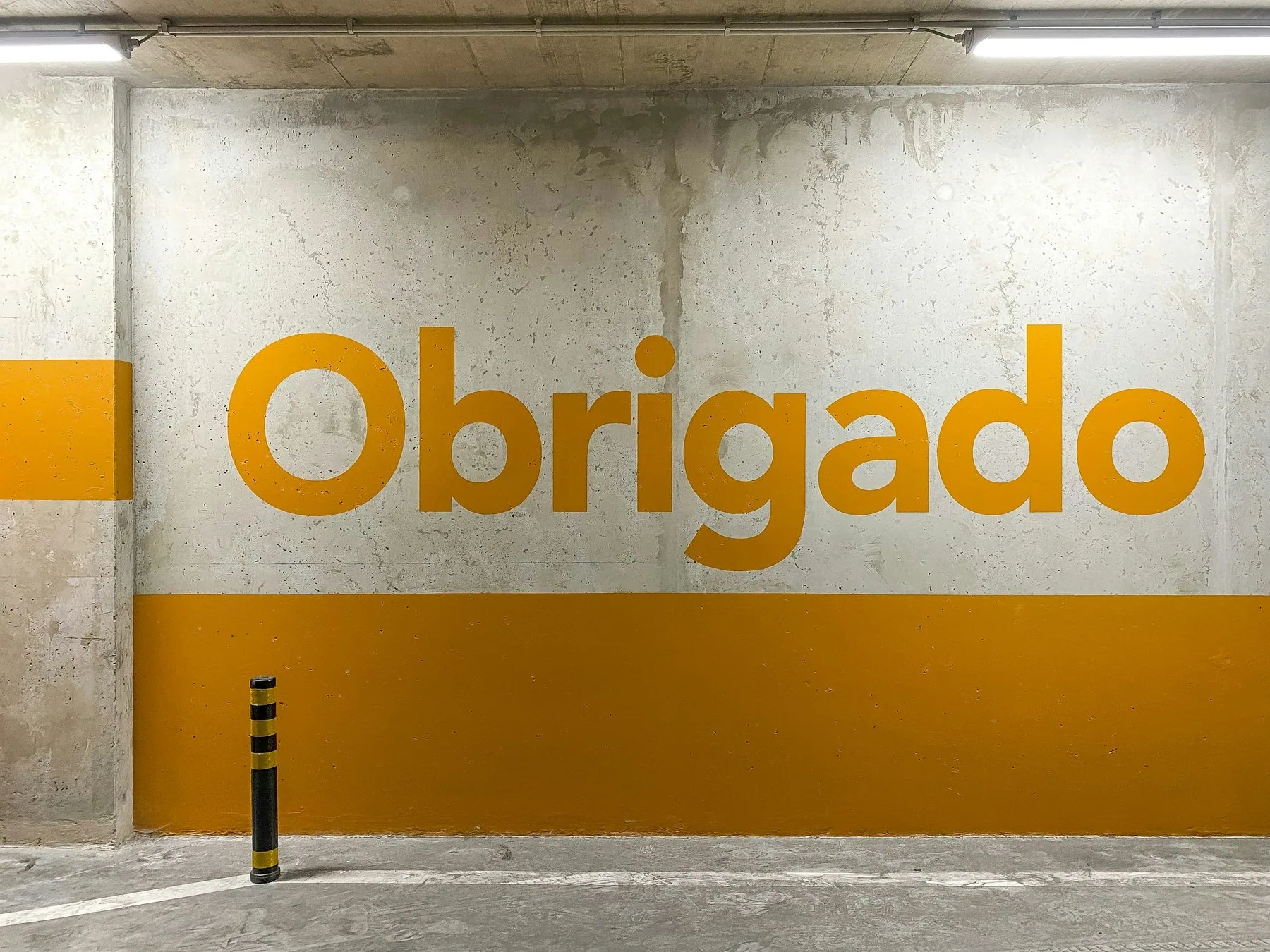 Det portugisiske ordet «Obrigado» malt med store gule bokstaver på en grå betongvegg med gul stripe