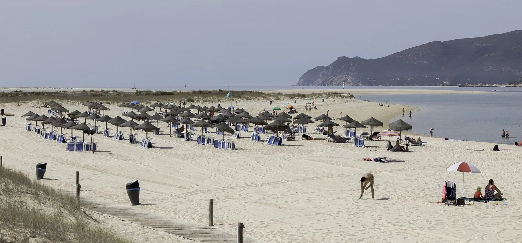 Bred sandstrand på Tróia med parasoller og få badegjester, og Arrábida-fjellene over Sado-munningen