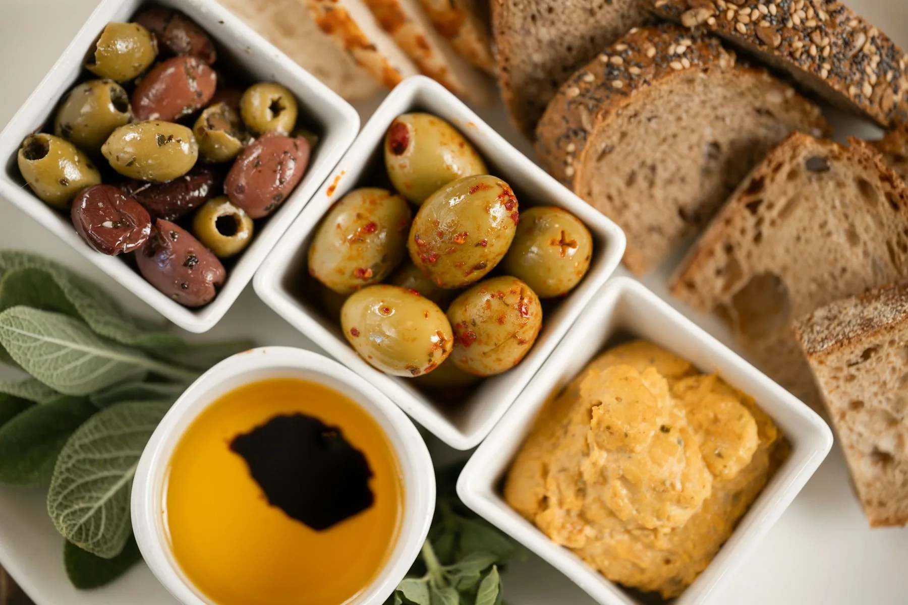 Små skåler med oliven, olivenolje og pålegg ved siden av brødskiver på et restaurantbord