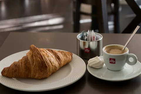 Croissant på hvit tallerken ved siden av en kopp kaffe og en liten marengs på et kafébord i Porto