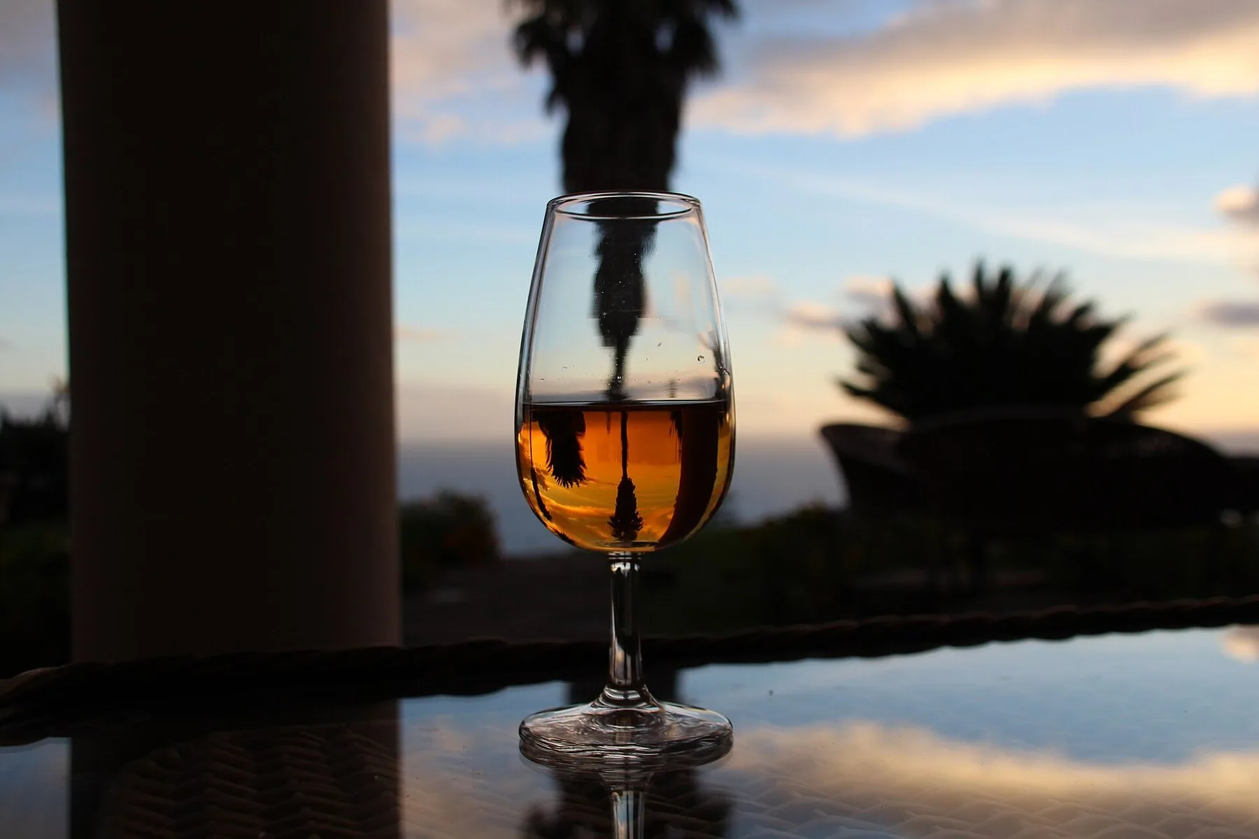 Et stettglass med ravfarget madeiravin på et bord i solnedgang, med palmer og havet i bakgrunnen i Calheta