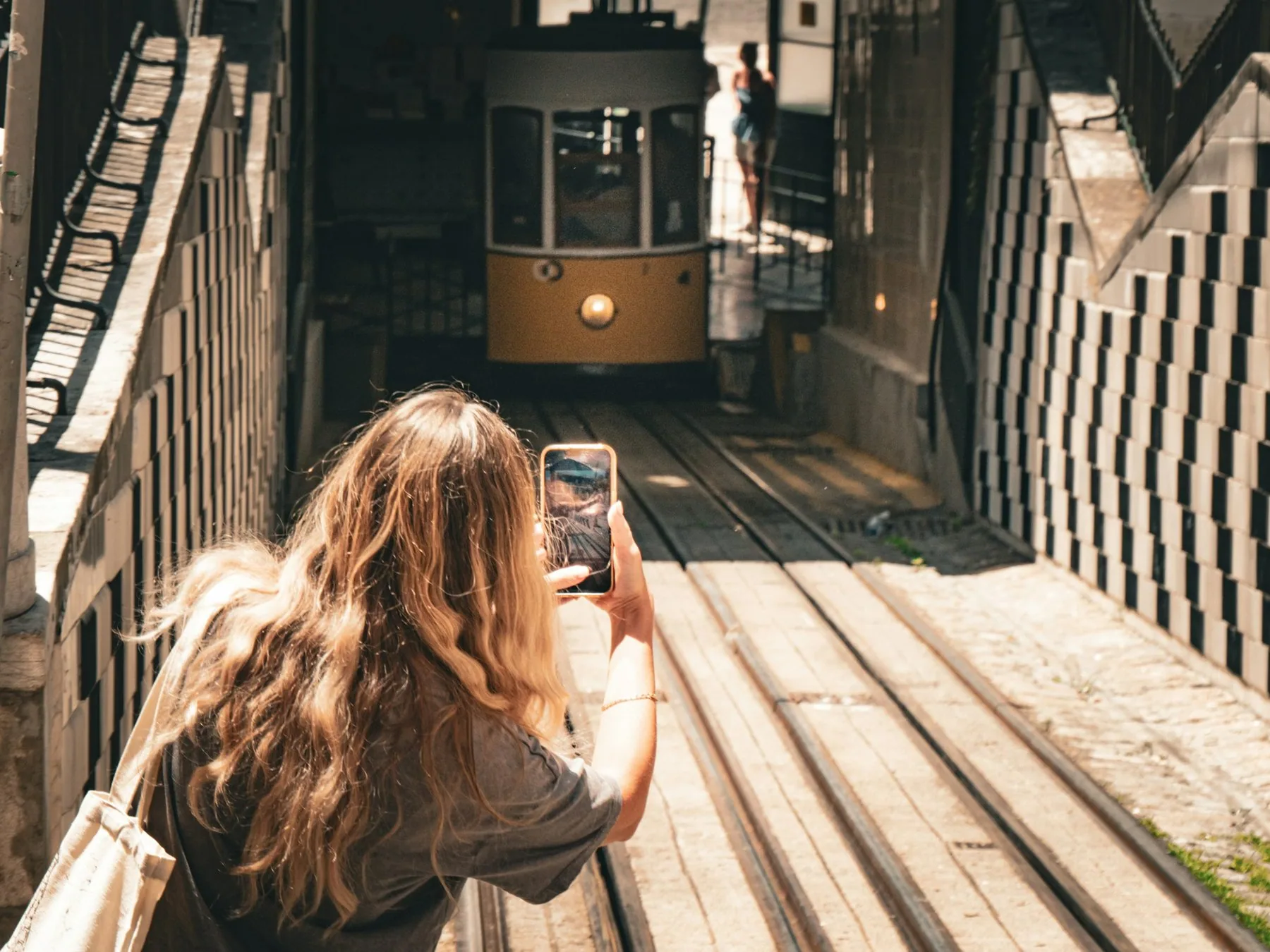 Kvinne sett bakfra tar bilde med mobilen av en gul kabelbane i en flislagt gate i Lisboa