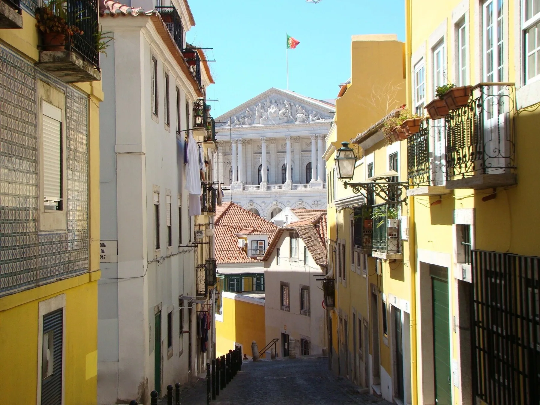 Smal brosteinsgate med gule hus og balkonger som leder mot parlamentsbygningen São Bento i Lisboa