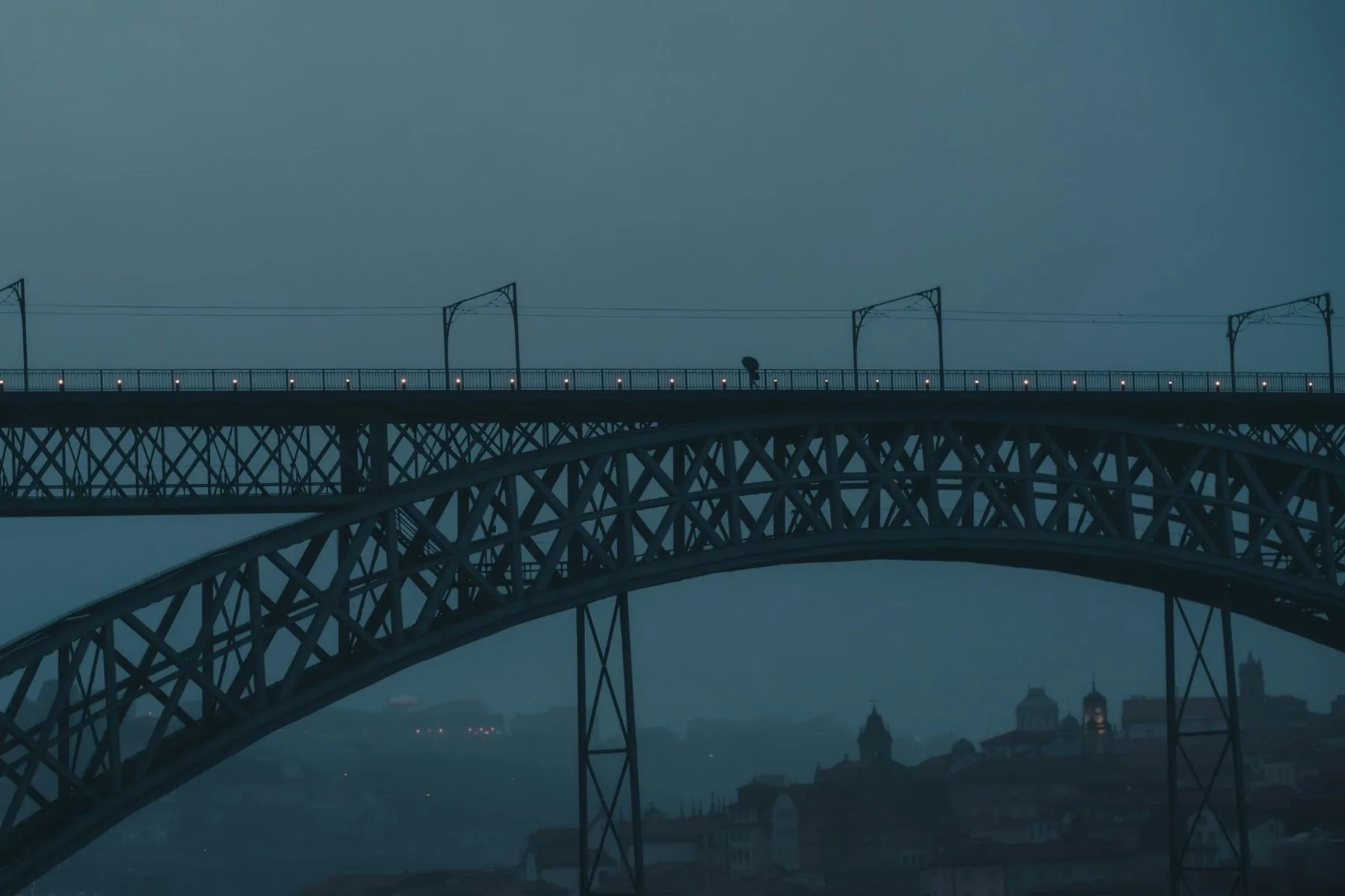 Jernbuen på Dom Luís I-broen i tåke og regnvær, med en liten skikkelse under paraply og silhuetter av Porto bak