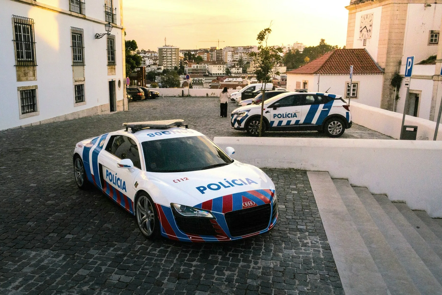 To hvite politibiler med blå og røde striper parkert på en brosteinsplass i Leiria i kveldslys