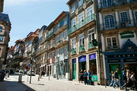 Solfylt brosteinsgate i Porto med høye, smale hus og et apotek med grønt skilt og blå fasade på hjørnet