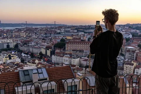 En mann fotografert bakfra holder opp mobilen mot utsikten over Lisboa fra et utsiktspunkt