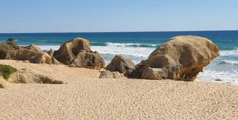 Avrundede sandsteinsblokker på en solfylt sandstrand der bølgene bryter inn fra Atlanterhavet i Algarve