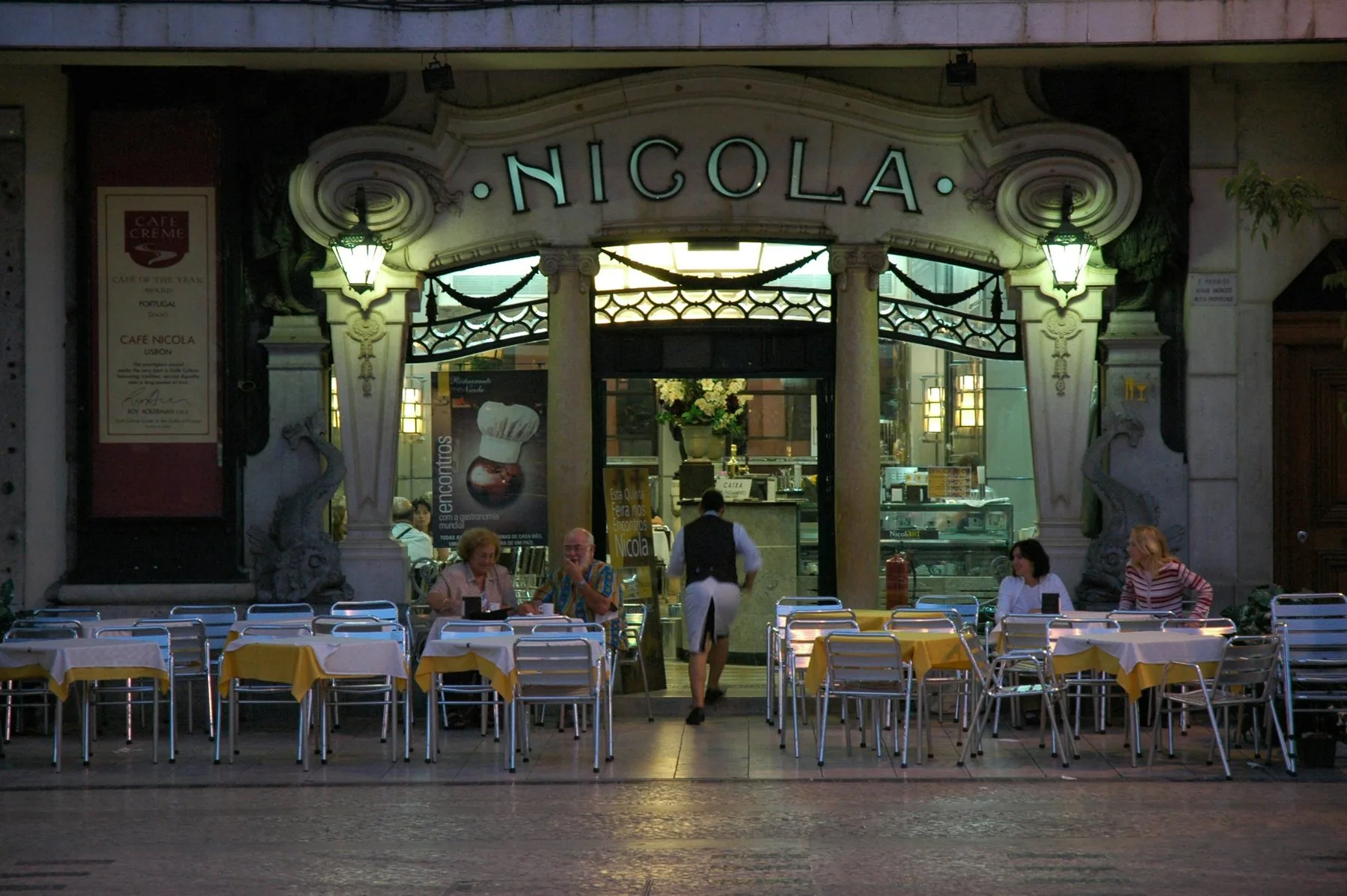 Uteserveringen foran art deco-fasaden til Café Nicola på Rossio i Lisboa i kveldslys, med gule duker og en kelner