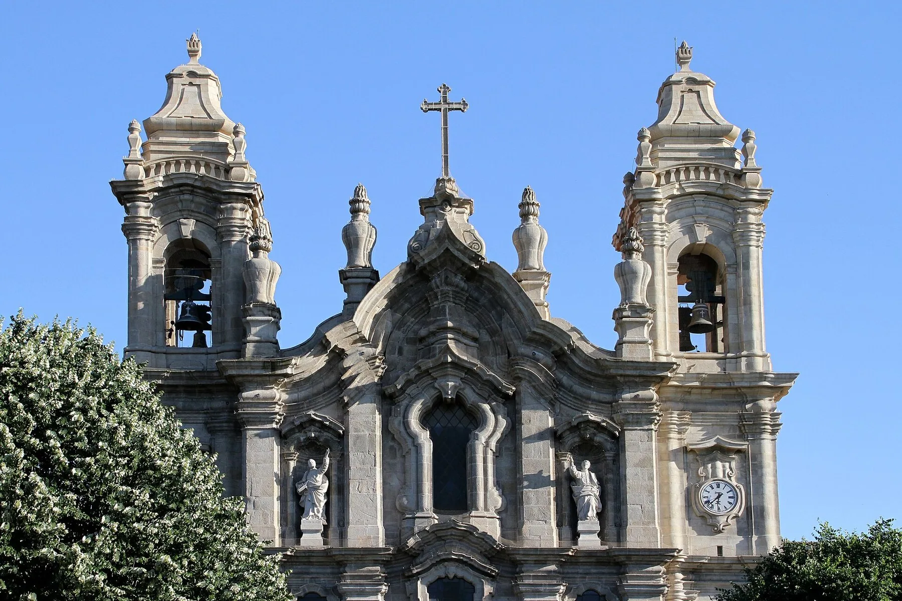 Barokkfasaden på Igreja dos Congregados i Braga med to klokketårn, helgenstatuer og blomstrende tre mot blå himmel