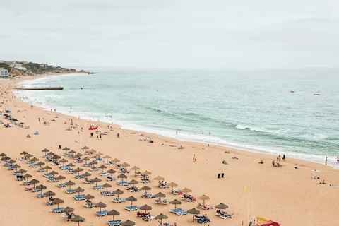 Lang sandstrand i Albufeira med rekker av stråparasoller og solsenger langs en rolig, grønnblå sjø