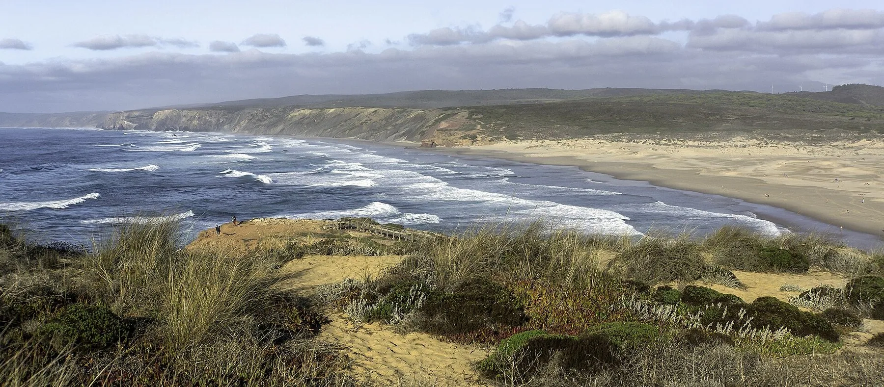 Bred sandstrand med hvite bølger og gressbevokste dyner i forgrunnen ved Praia da Bordeira nær Carrapateira