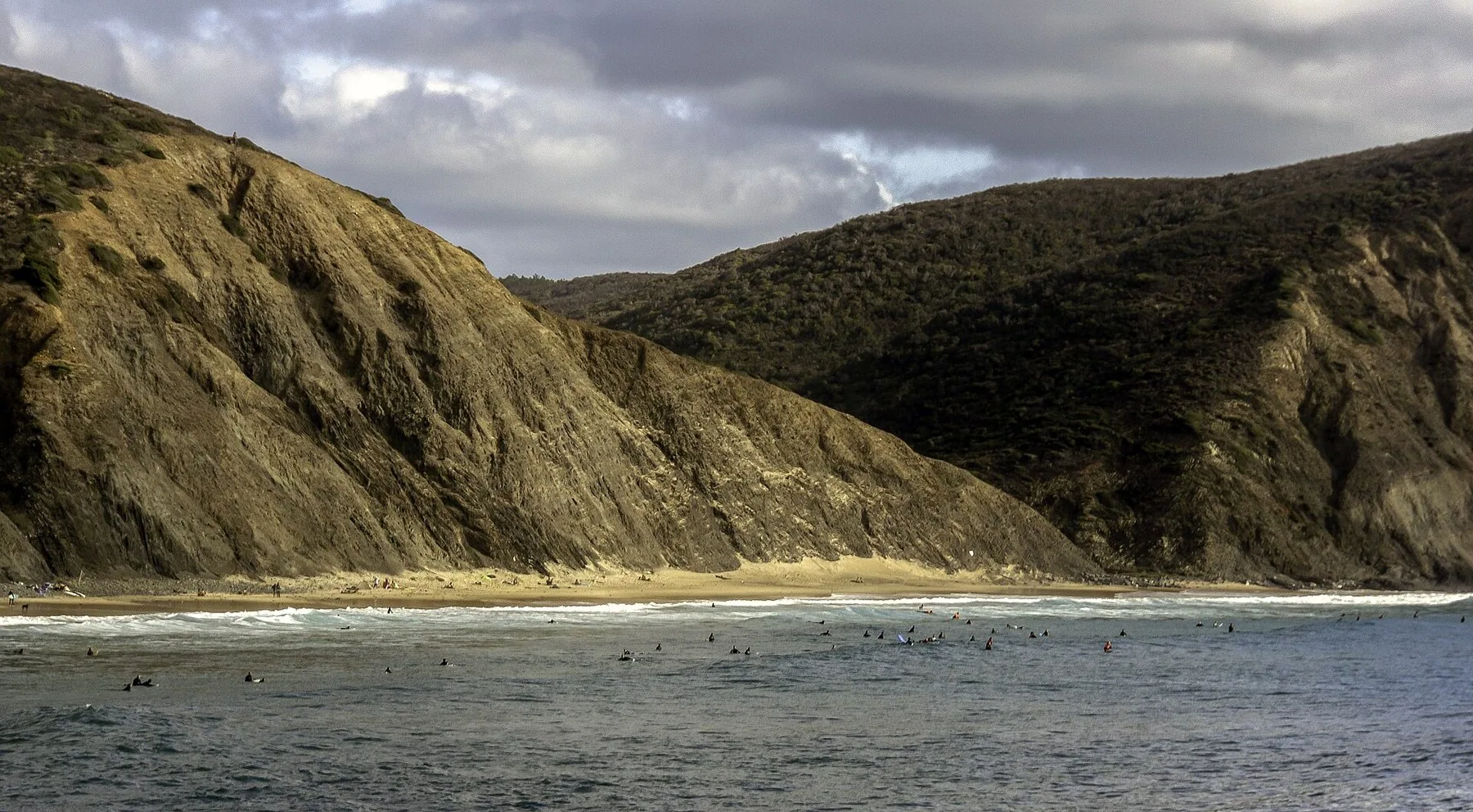 Surfere venter på bølgene i bukta ved Praia da Arrifana under bratte, brune åser og en grå himmel