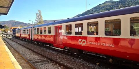 Rødt og hvitt regionaltog står ved perrongen på Pinhão stasjon med grønne åser i Dourodalen bak