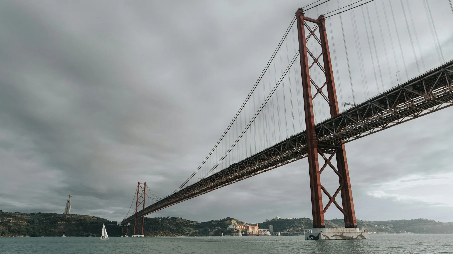 Den røde hengebrøn 25 de Abril spenner over Tejo mot sørbredden og Cristo Rei under grå skyer