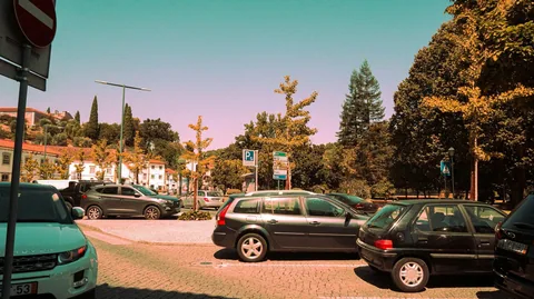 Biler parkert på en brosteinsbelagt plass med blått P-skilt, trær og hvite hus i bakgrunnen