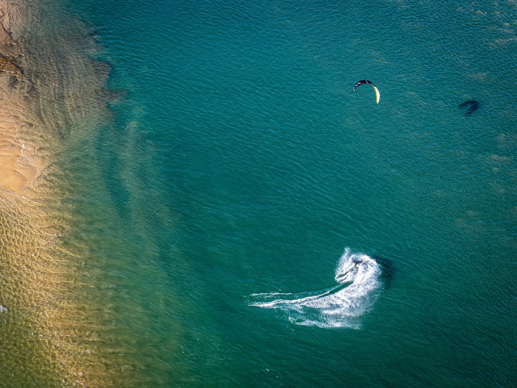 Kitesurfer drar en hvit skumstripe over grønnblått vann ved en sandstrand utenfor Sines, sett ovenfra